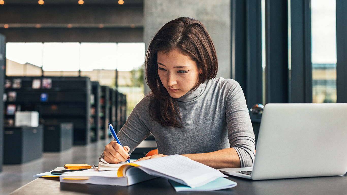 Woman with a laptop taking notes while referencing an open book