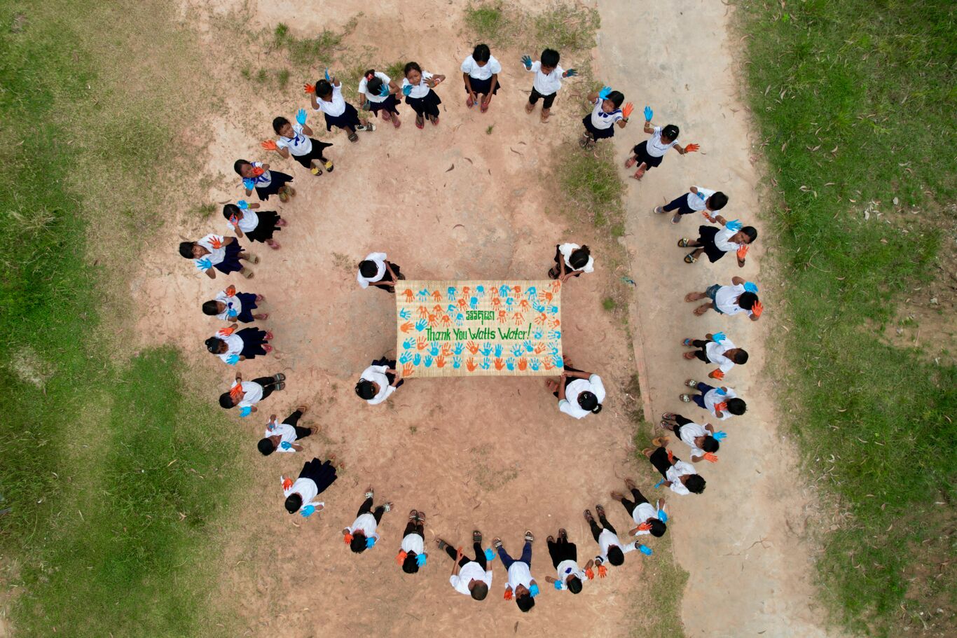 Aerial view of a circle of children, with their hands painted and pointed towards the sky. Circling 4 children holding a Thank You banner.